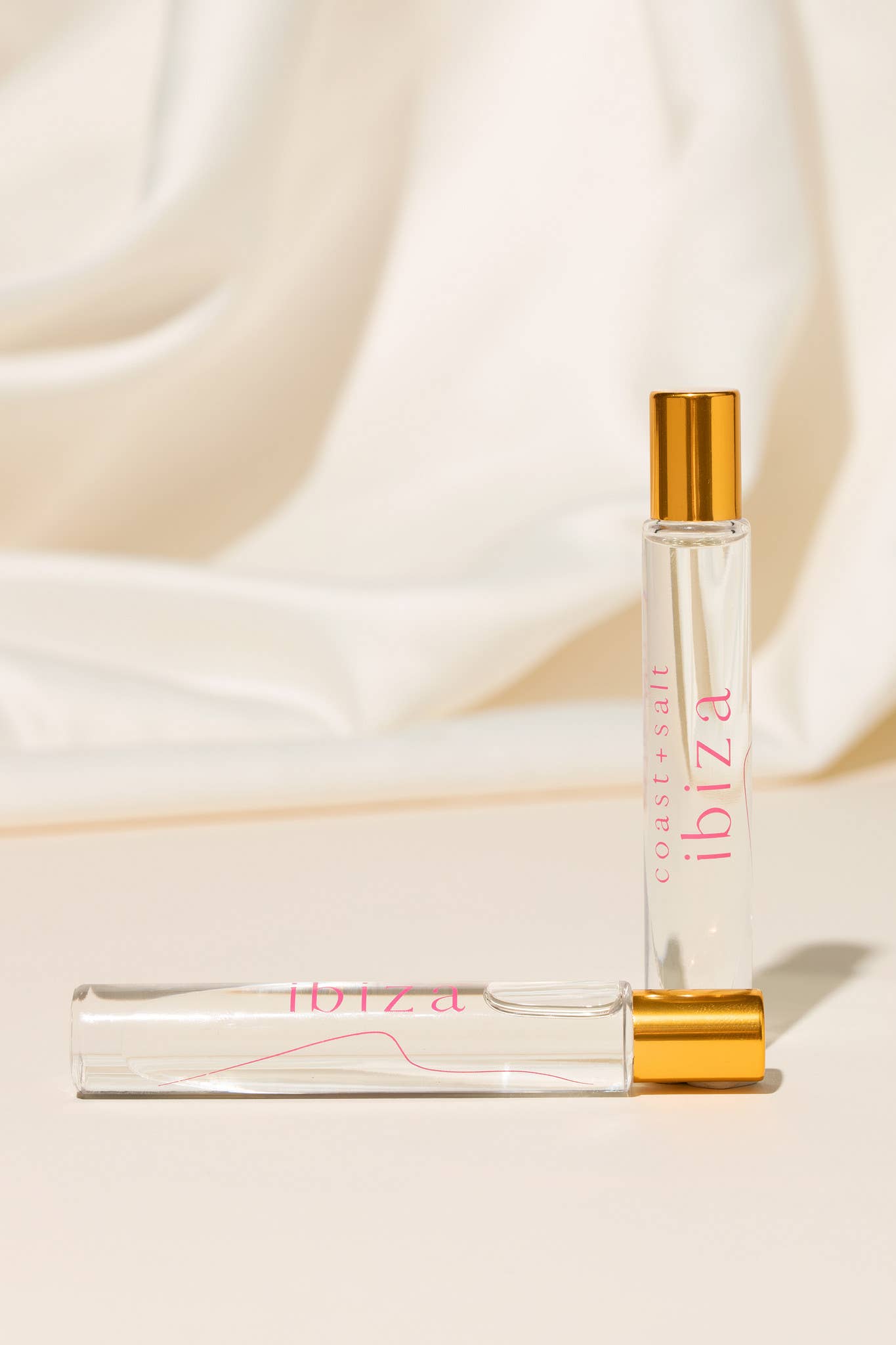 Two perfume rollers with gold caps rest on a cream surface; both are compact and designed for on-the-go fragrance use.