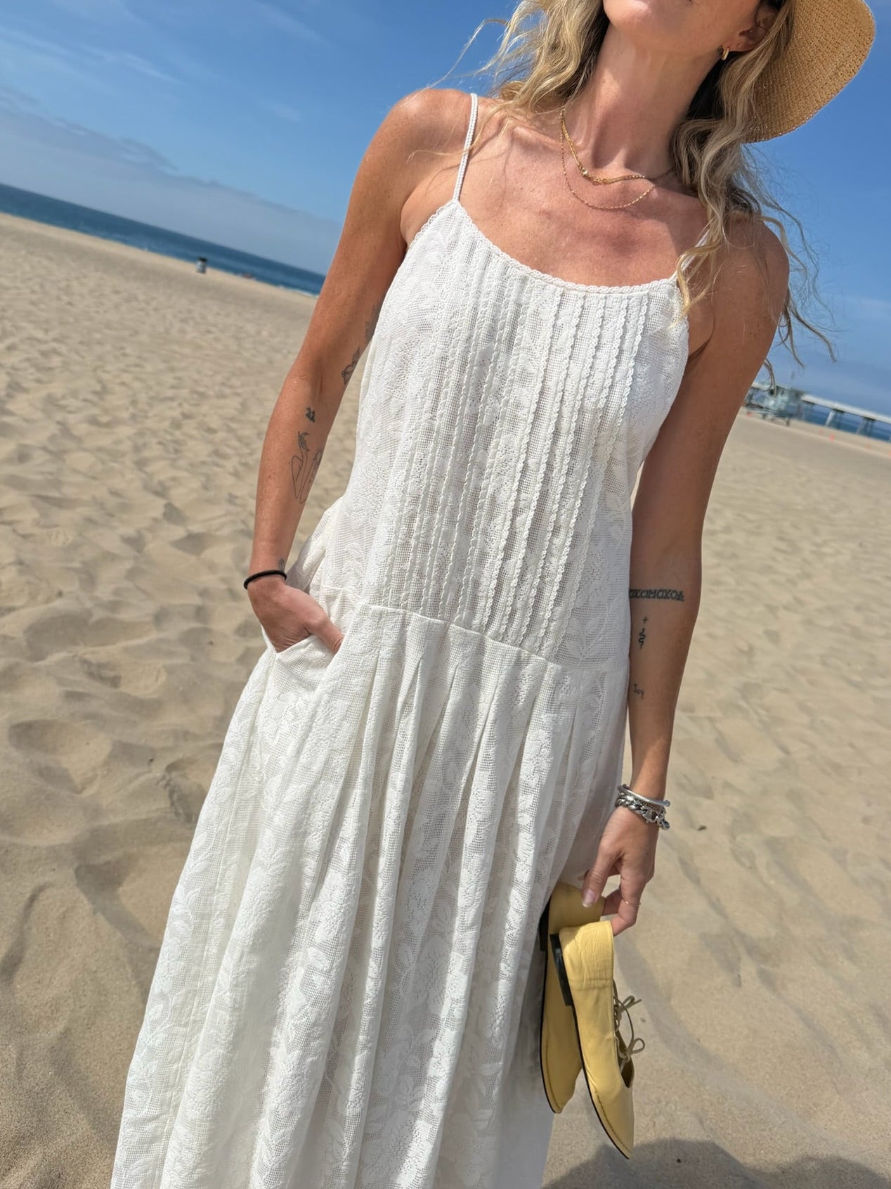 Woman in a white midi dress and straw hat stands barefoot on sandy beach, holding yellow shoes.