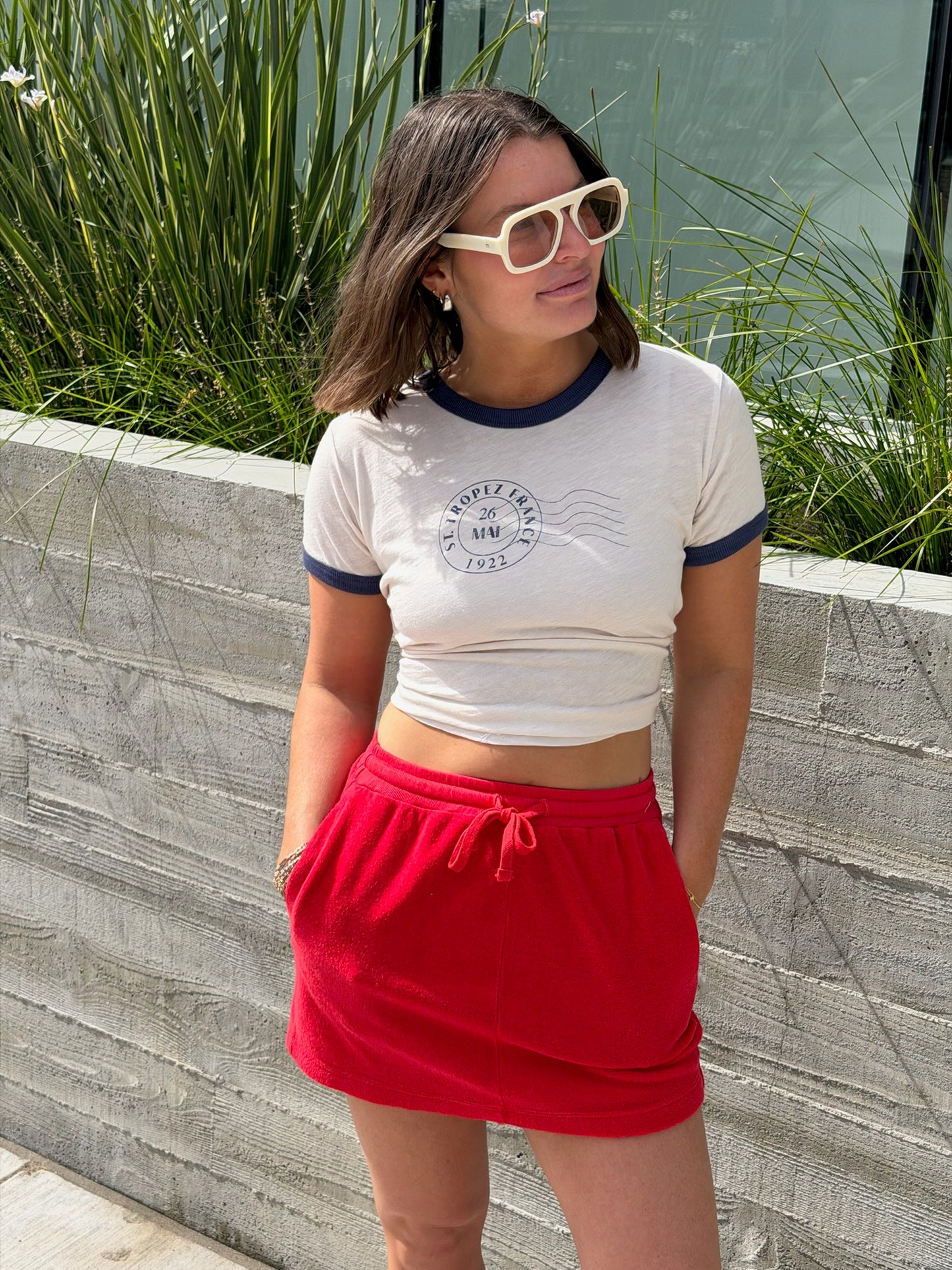 Woman in white sunglasses and crop top wears a vermillion terry mini skirt, standing by a concrete wall and tall grass.
