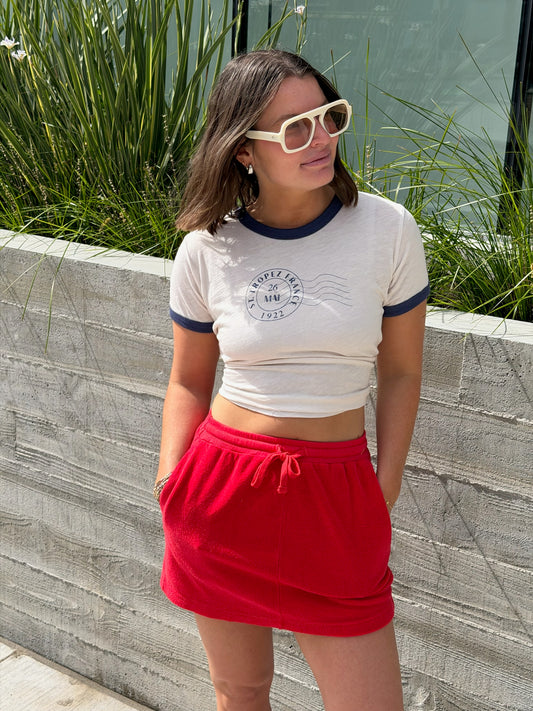 Woman in white sunglasses and crop top wears a vermillion terry mini skirt, standing by a concrete wall and tall grass.