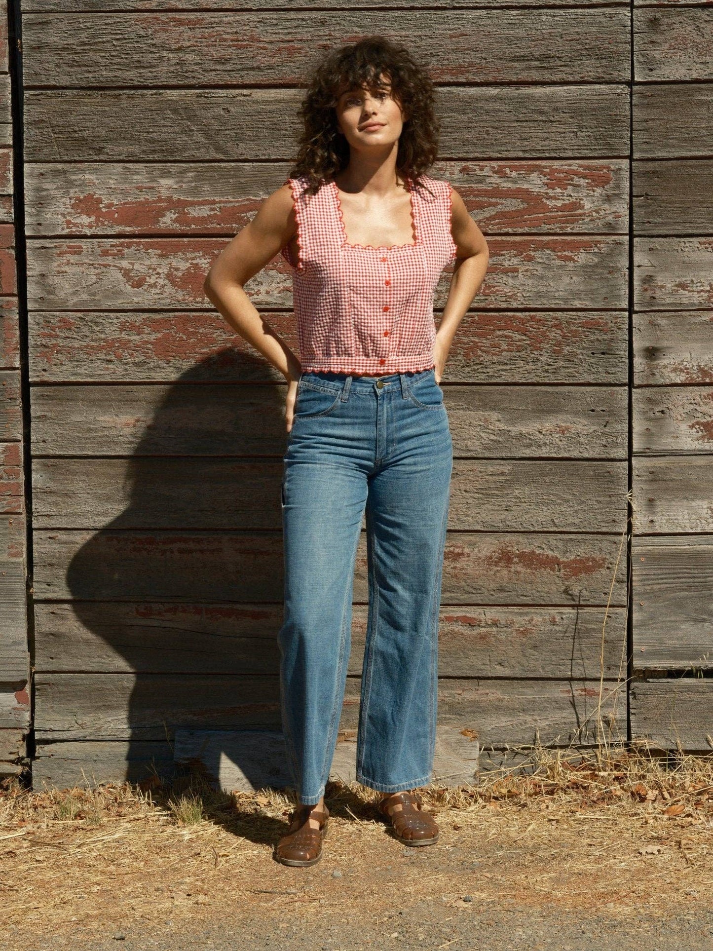 Woman wears a sleeveless cotton top and blue jeans, standing by an aged wooden wall outdoors in sunlight.