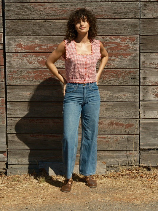 Woman wears a sleeveless cotton top and blue jeans, standing by an aged wooden wall outdoors in sunlight.
