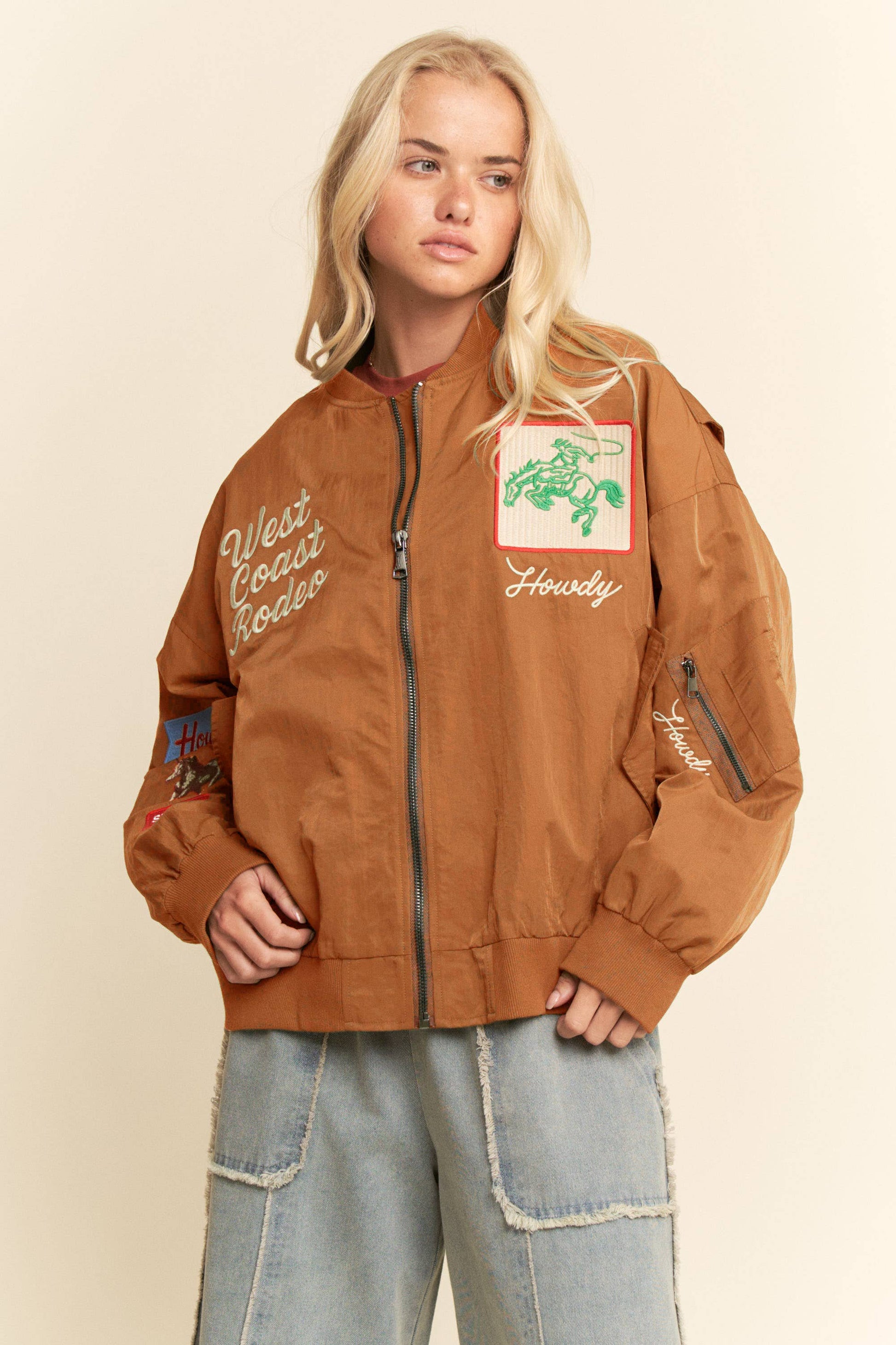 Woman wearing embroidered bomber jacket and light denim jeans, standing before a beige background.