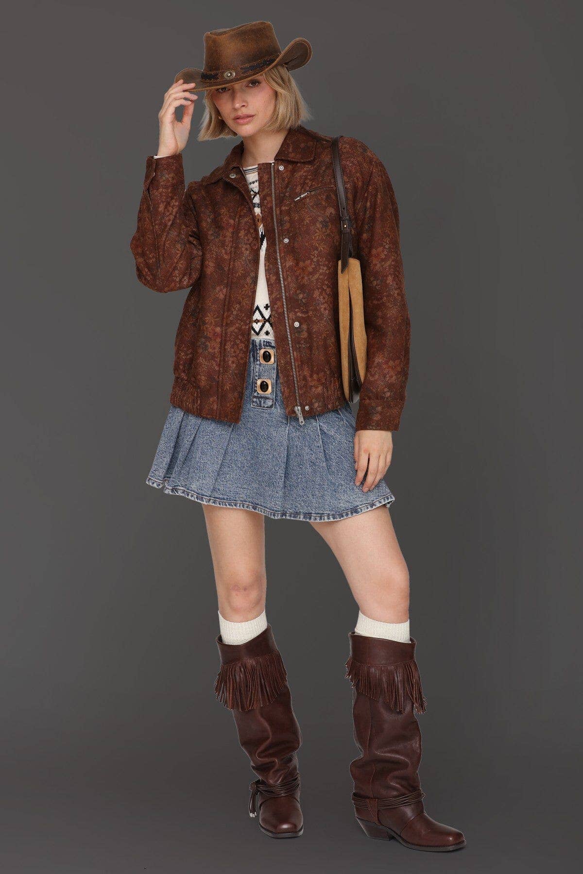 Woman in brown hat and fringe boots wearing a pleated denim skirt and floral print bomber jacket.