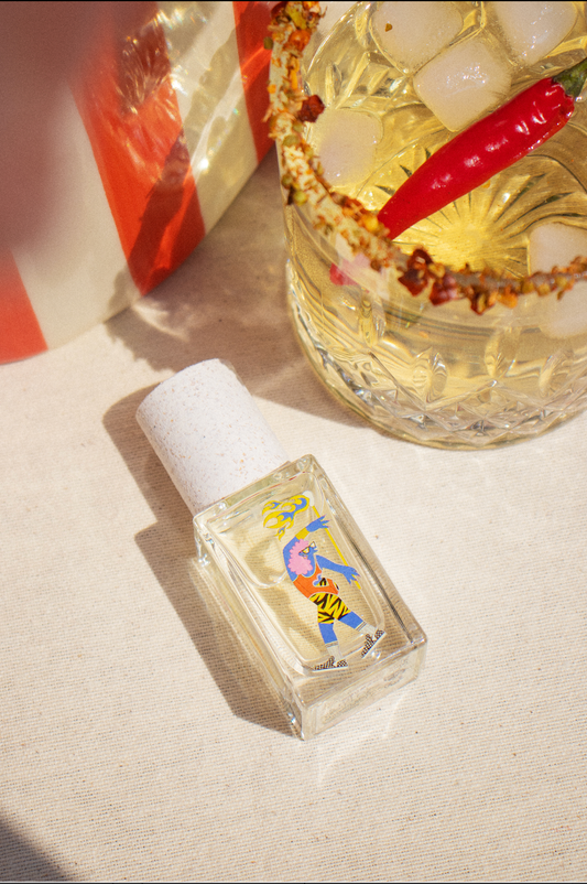 A small glass perfume bottle rests next to an iced beverage with red chili and spice garnish.