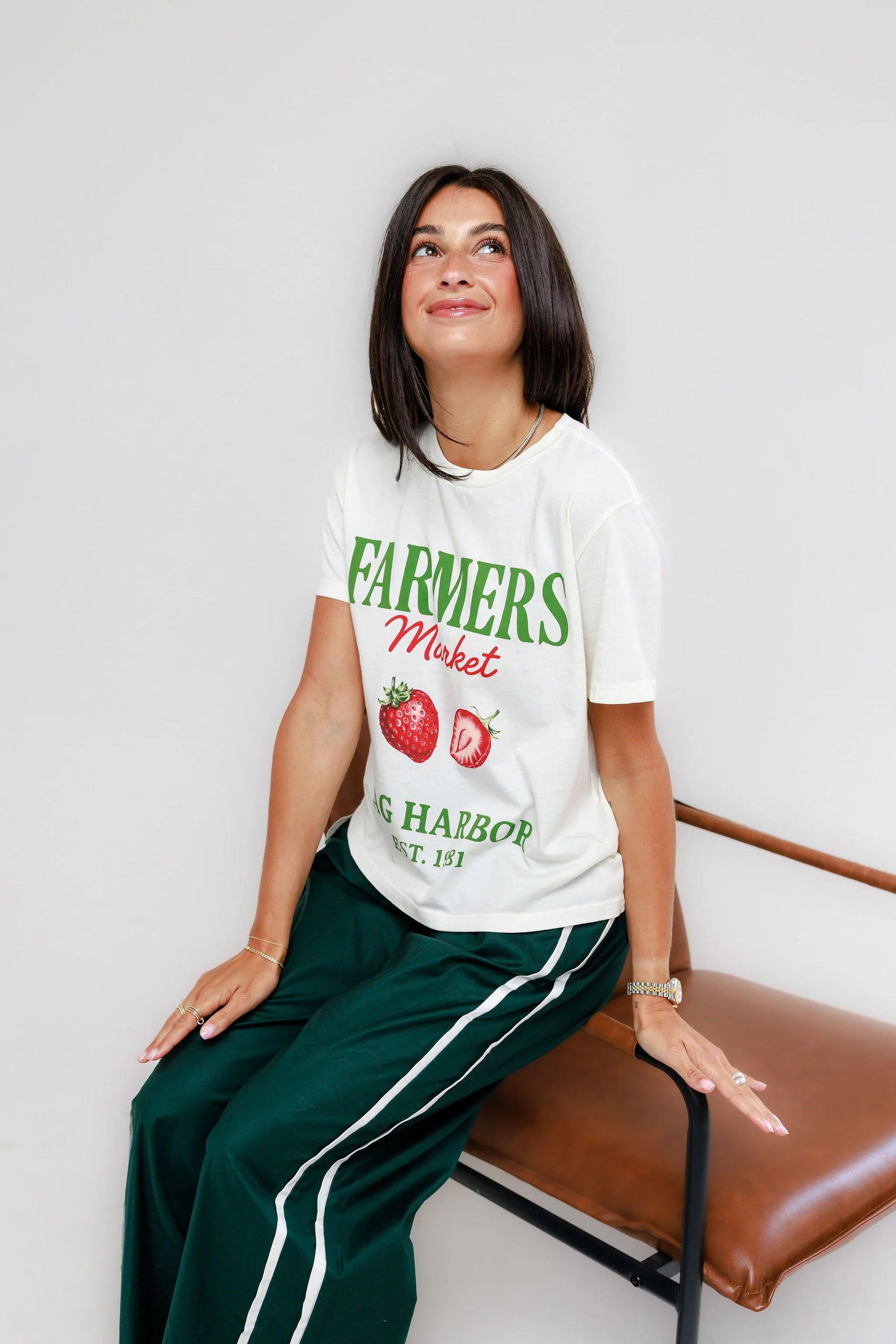 Woman with dark hair sits on a brown chair, wearing an off-white cotton tee and green pants.