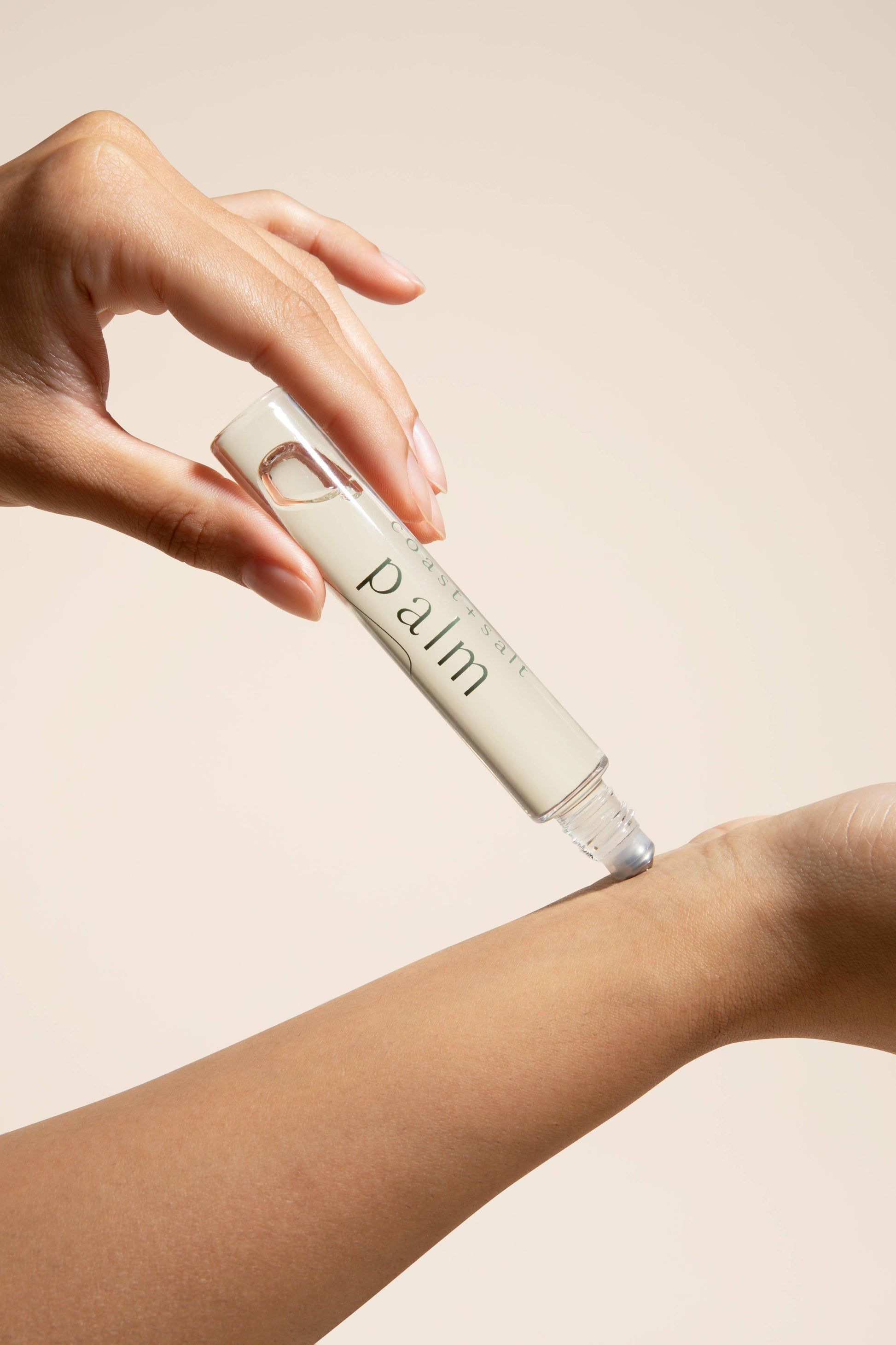 A hand rolls a clear glass perfume applicator onto the inner wrist, with a neutral background behind.