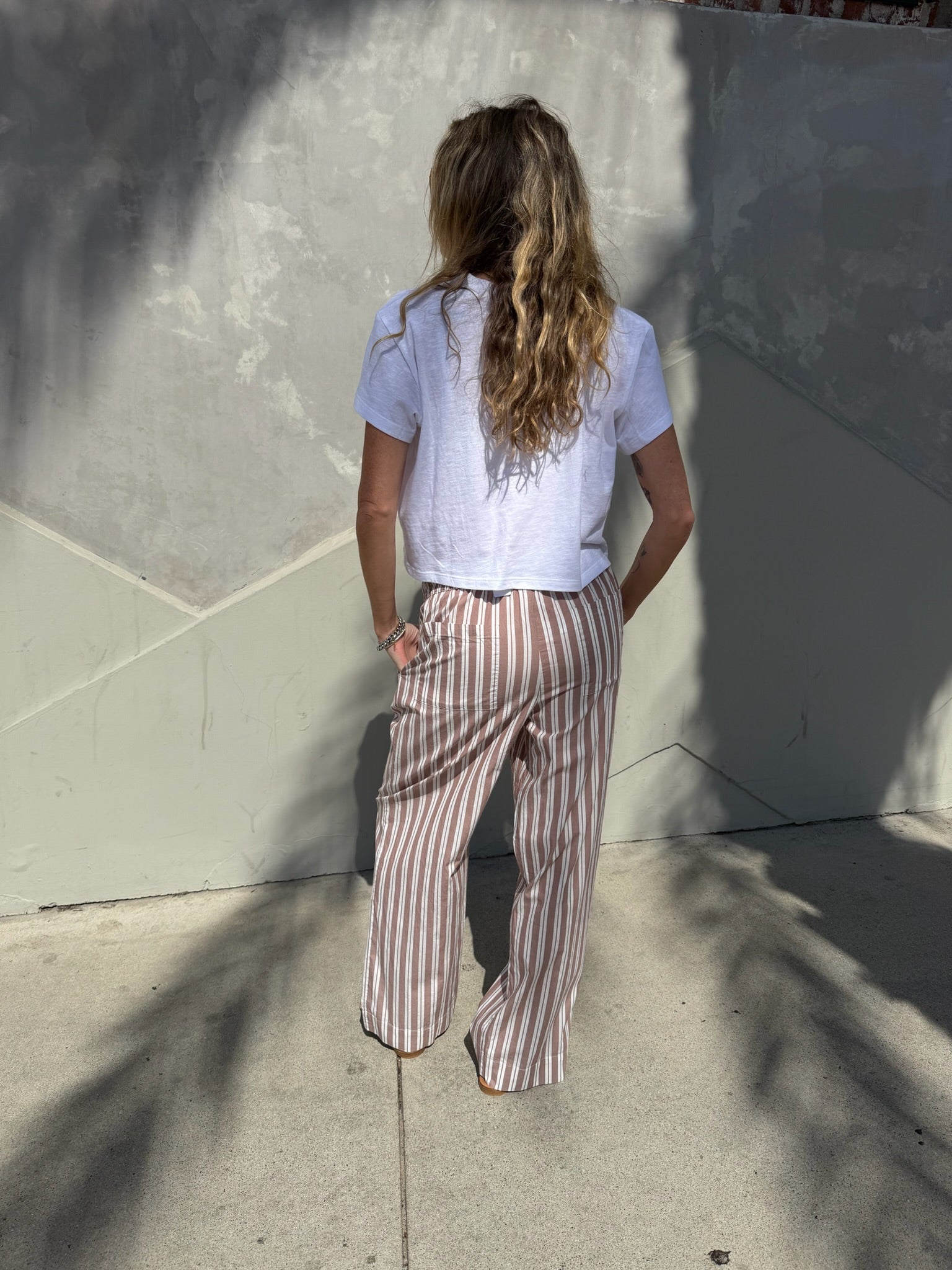 Woman with long wavy hair in beige striped wide-leg lounge pants and a white shirt stands near a sunlit concrete wall.