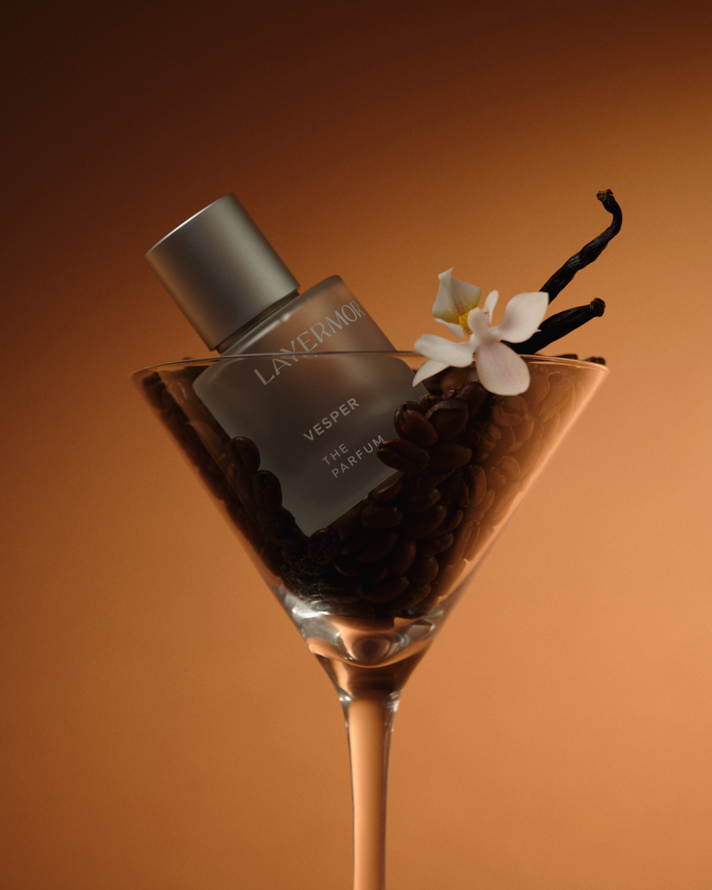Body oil displayed in a clear martini glass, surrounded by coffee beans, vanilla pods, and a white flower.