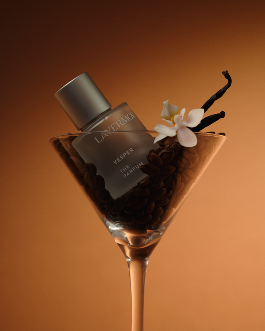 Body oil displayed in a clear martini glass, surrounded by coffee beans, vanilla pods, and a white flower.