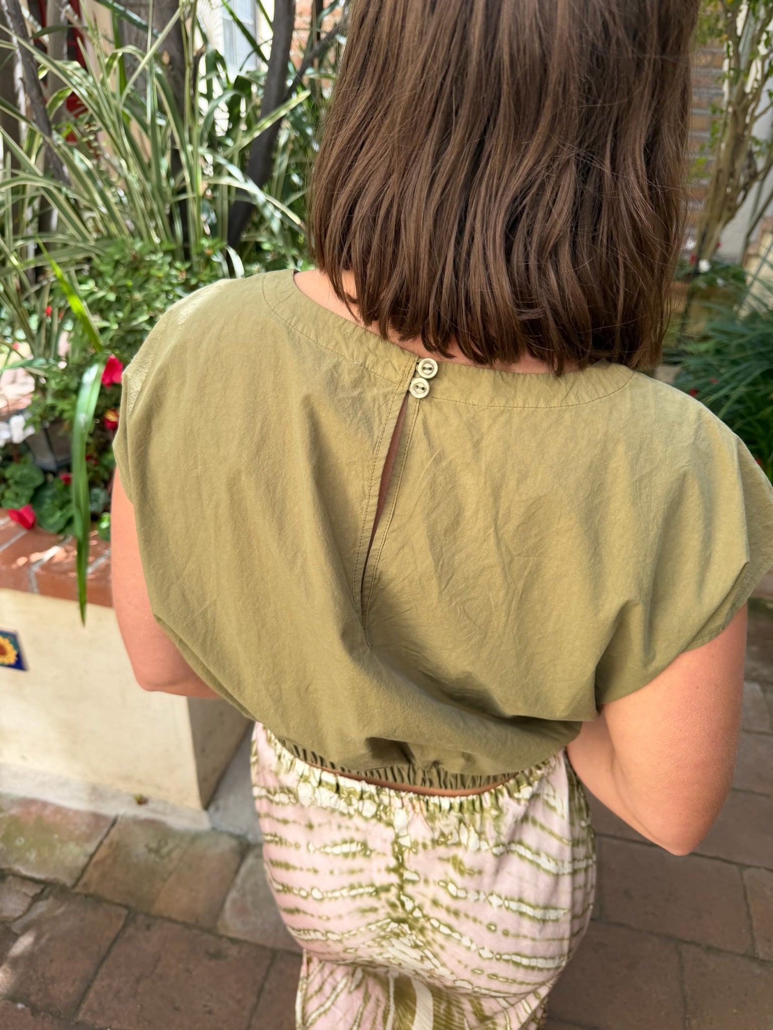Woman with shoulder-length brown hair wears a dark olive top outdoors near green plants.