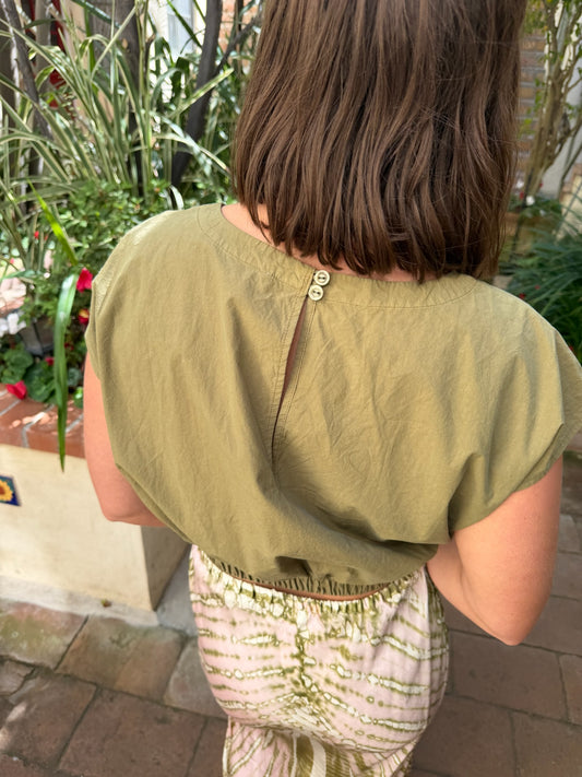 Woman with shoulder-length brown hair wears a dark olive top outdoors near green plants.