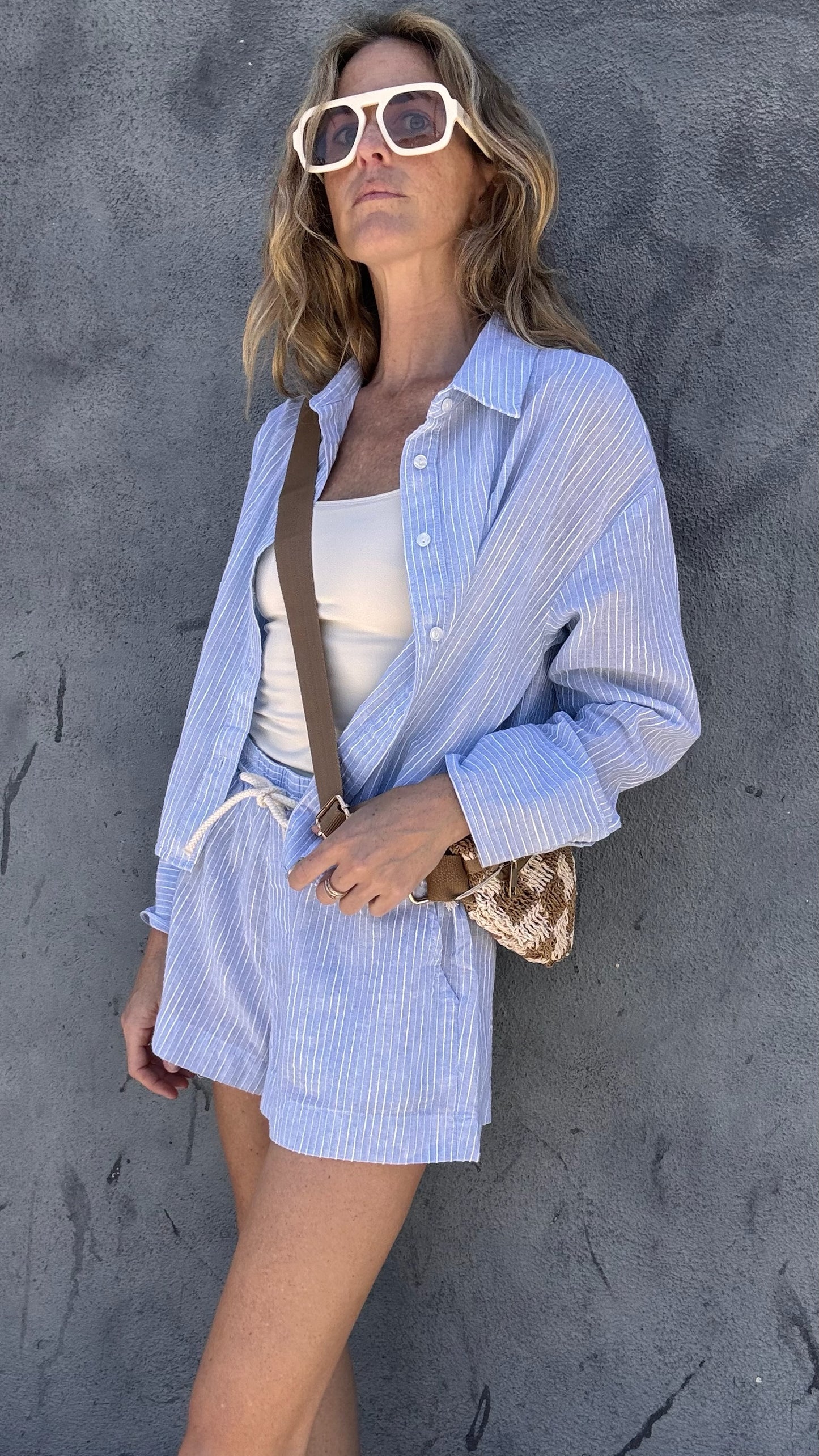 Woman in light blue button-up shirt over white tank with sunglasses and crossbody bag by a textured gray wall.