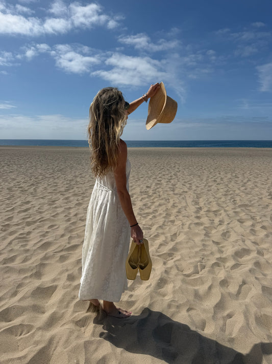 Woman barefoot on sandy beach in white cotton midi dress, holding shoes and raising hat overhead.