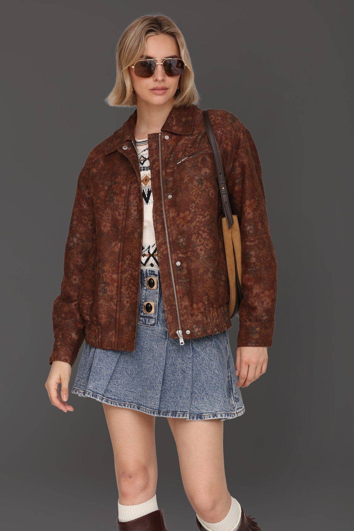 Woman wearing floral print bomber jacket, sunglasses, patterned sweater, denim skirt, and brown boots.