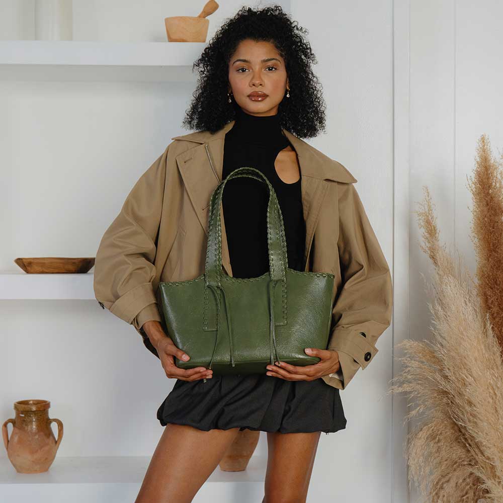 Woman in tan jacket holds a black recycled vegan leather shoulder bag while standing next to pottery shelves.