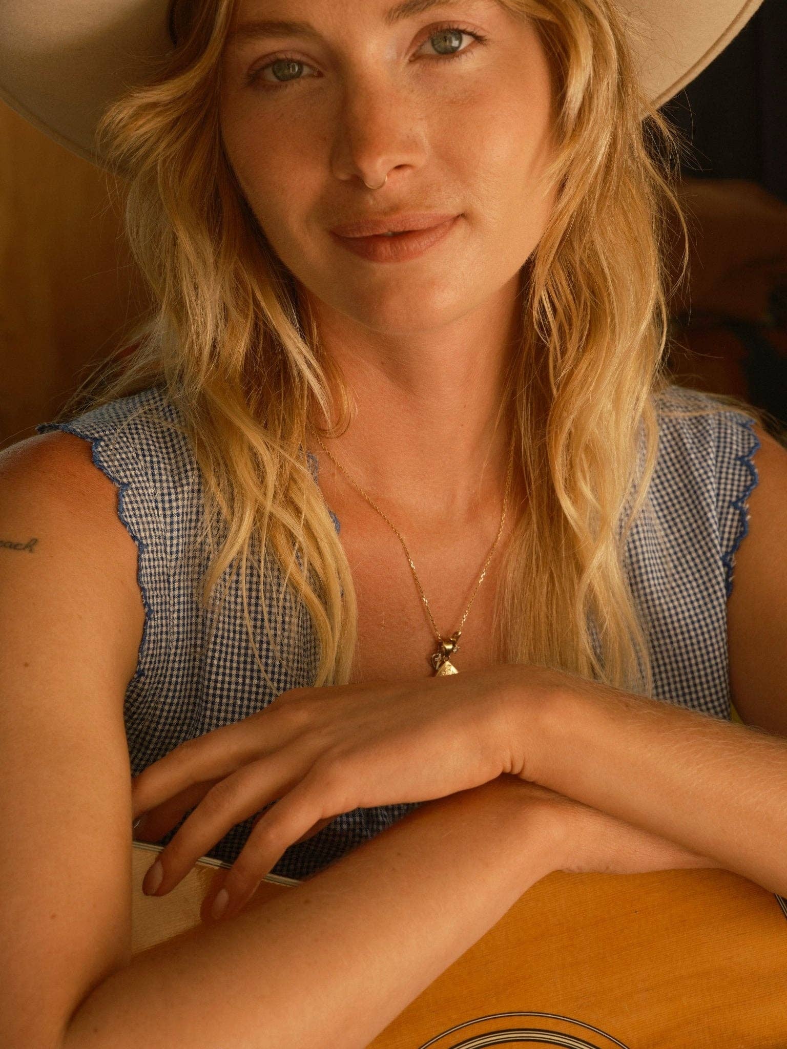 Woman wears a light fabric mini dress and hat, holding a guitar and smiling at the camera.