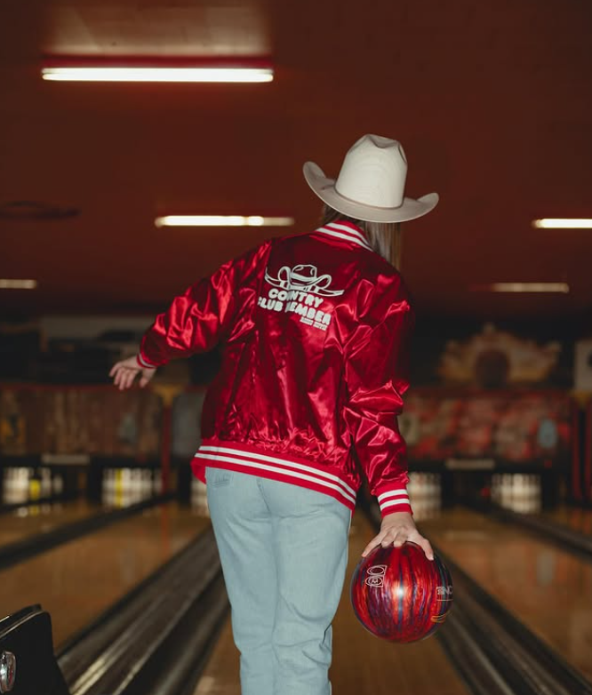 Person wearing a satin Rodeo Hippie COUNTRY CLUB MEMBER jacket and cowboy hat bowls with a red ball at the lane’s start.