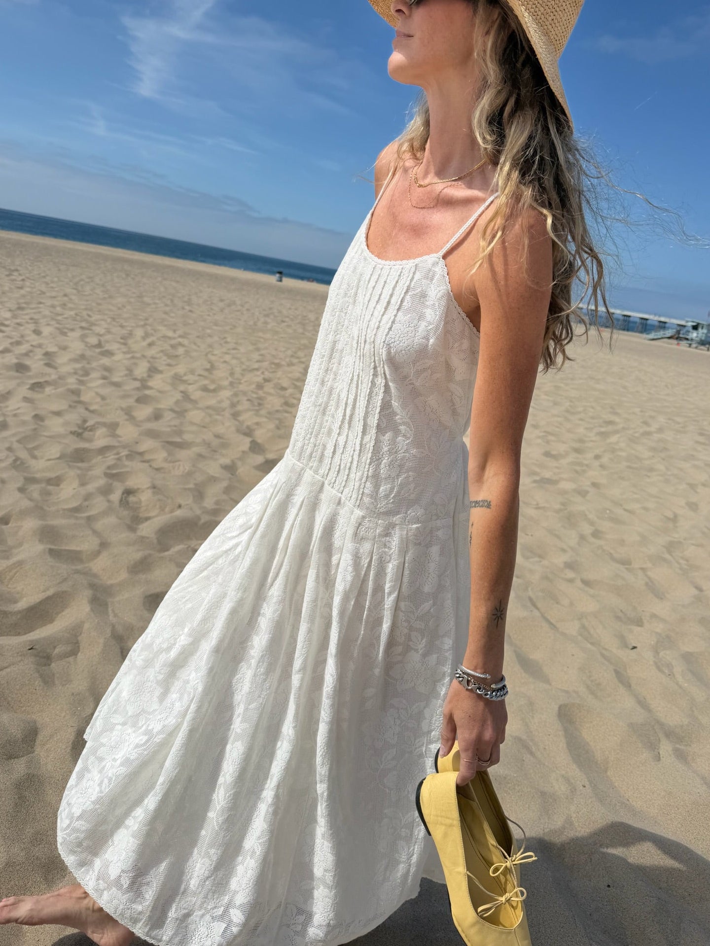 Woman in a white midi dress and straw hat walks barefoot on the beach beneath a clear blue sky.