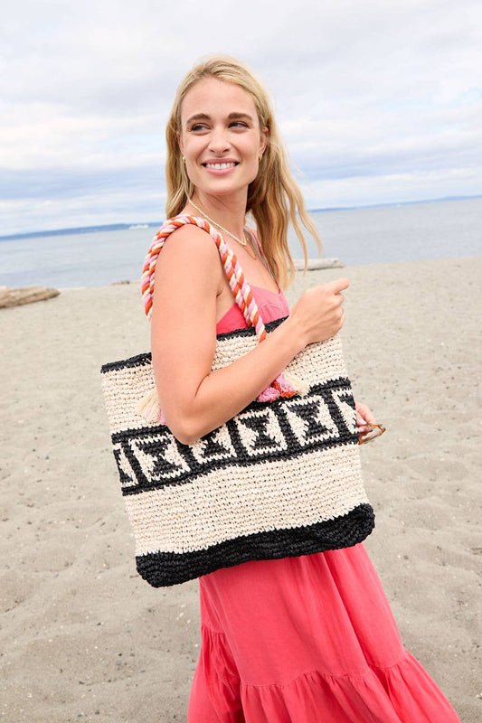 Woman in coral dress carries a woven Bandita tote bag on sandy beach by the ocean.