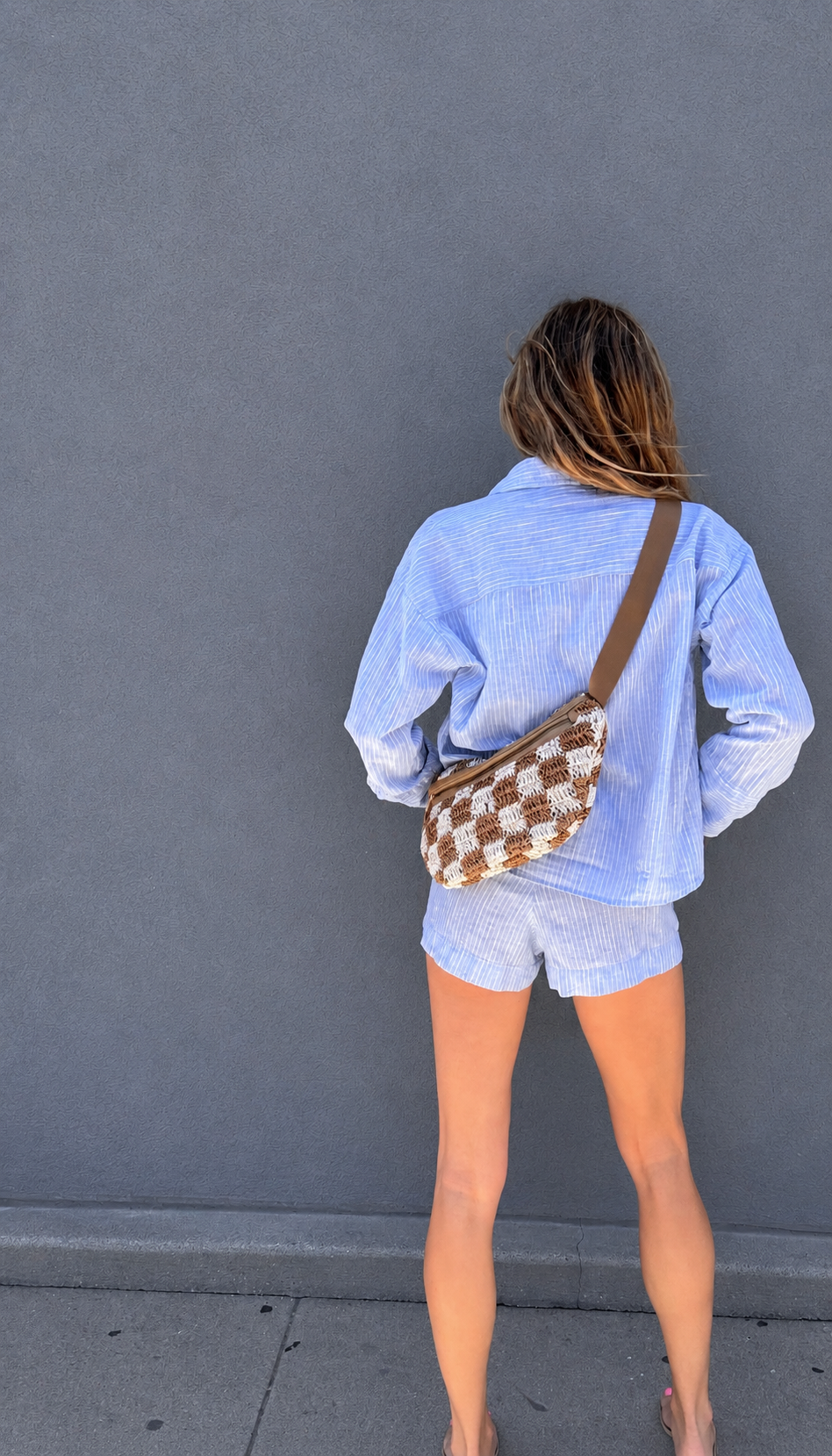 Woman in a light blue shirt with a brown and white checkered crossbody bag, standing facing a gray wall.