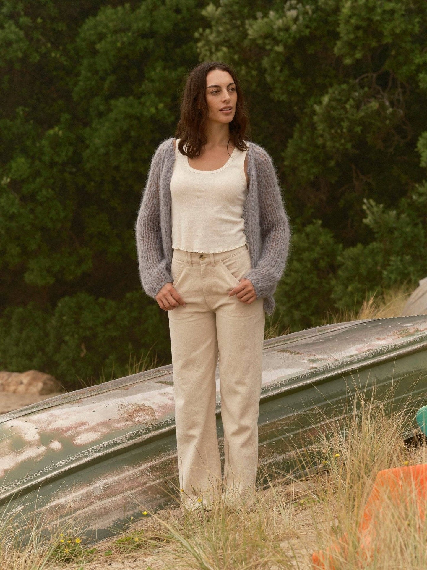 Woman in a light knit cardigan, beige pants, and white top stands outdoors near an overturned boat.