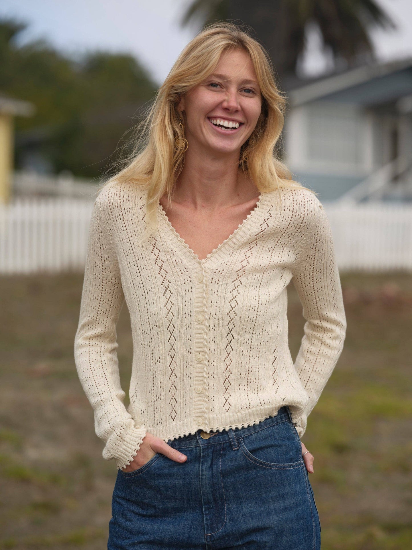 Woman outdoors in a knit cardigan and blue jeans; background features houses and a white fence.
