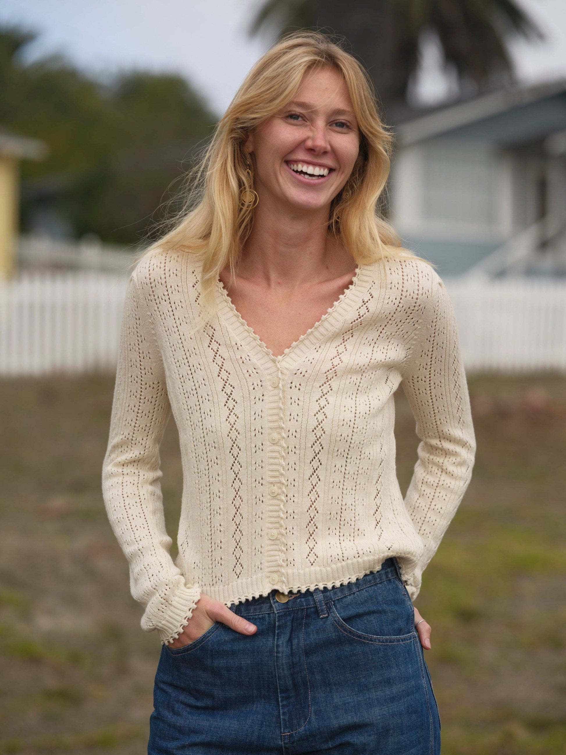 Woman outdoors in a knit cardigan and blue jeans; background features houses and a white fence.