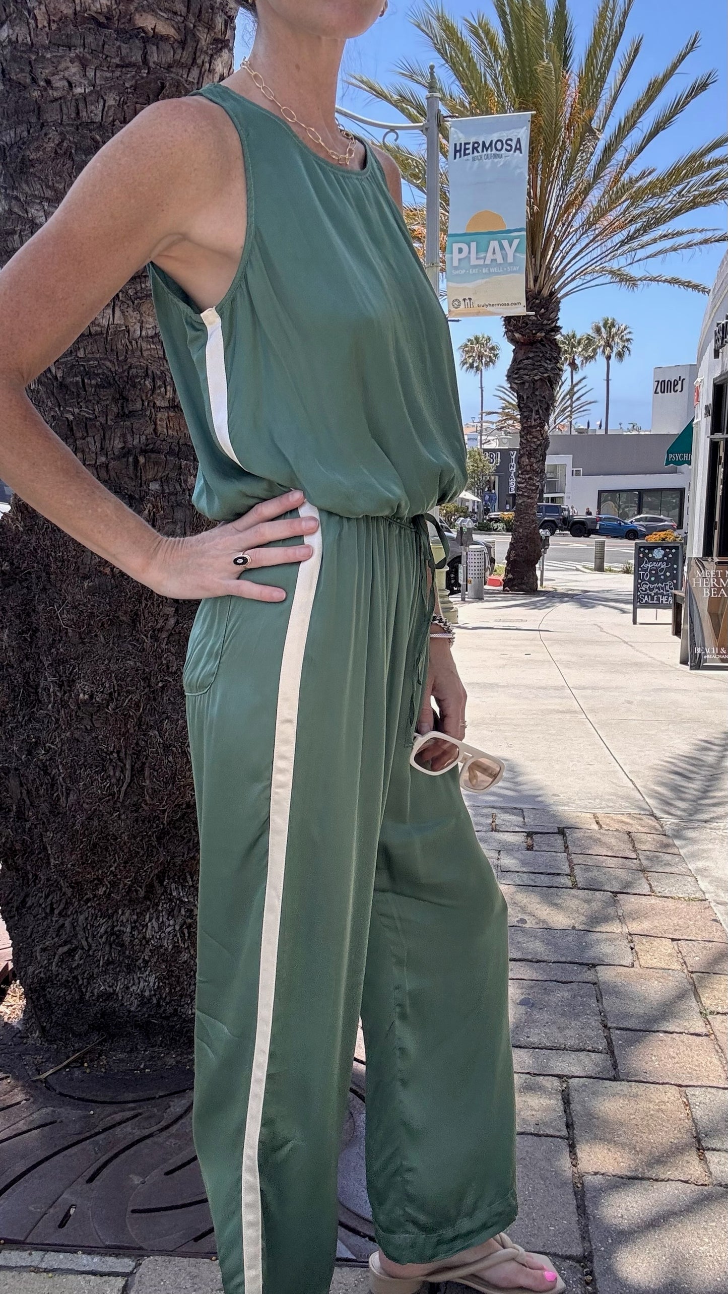Person stands on a sunny sidewalk with palm trees, holding sunglasses and wearing light green track pants.