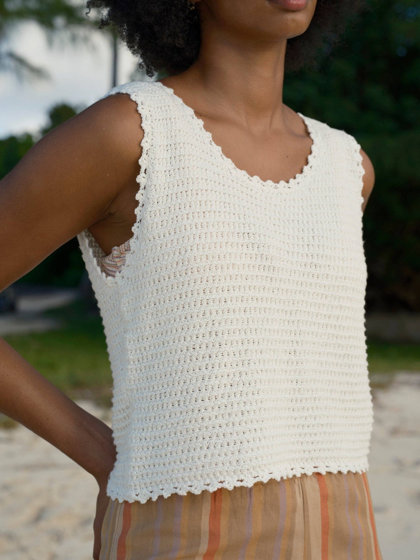 Woman wears a white crochet tank and striped skirt outdoors, with green foliage in the background.