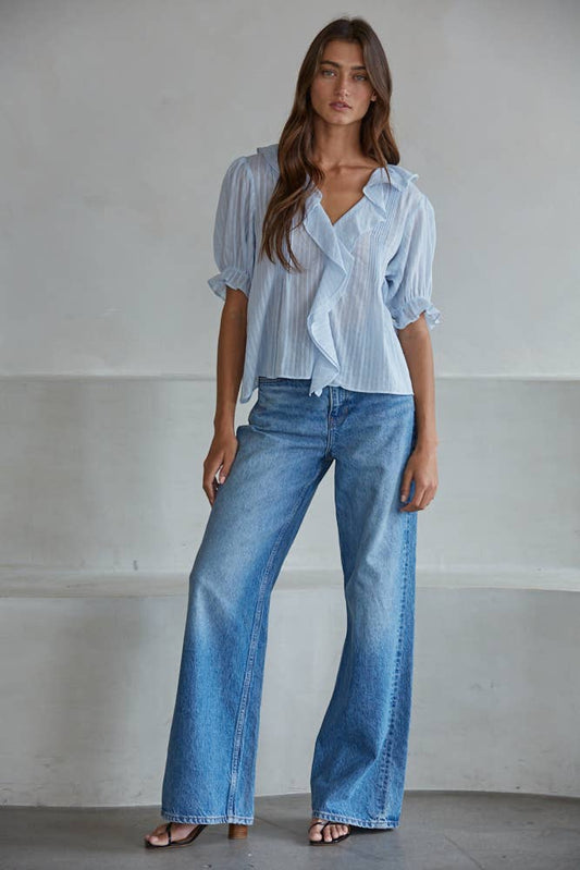 Woman wears powder blue top and wide-leg jeans, standing against a plain light wall. Cotton blend ideal for casual wear.