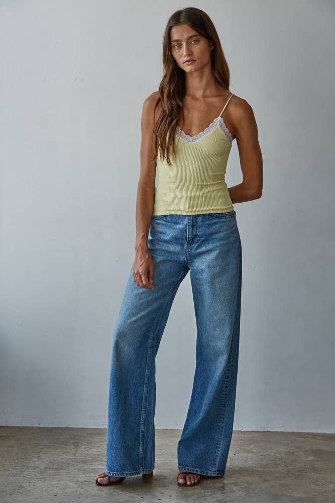 Woman in pastel yellow knit top and wide-leg jeans stands by a gray wall.