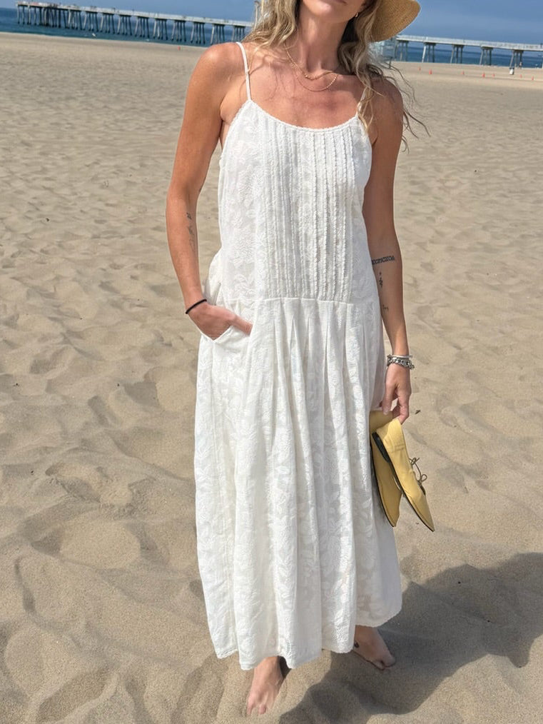 Woman in a white midi dress stands barefoot on the beach, holding sandals, with a pier visible in the background.