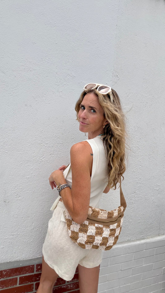 Woman with wavy blonde hair wears a parchment-colored open side top, sunglasses, and carries a woven bag.