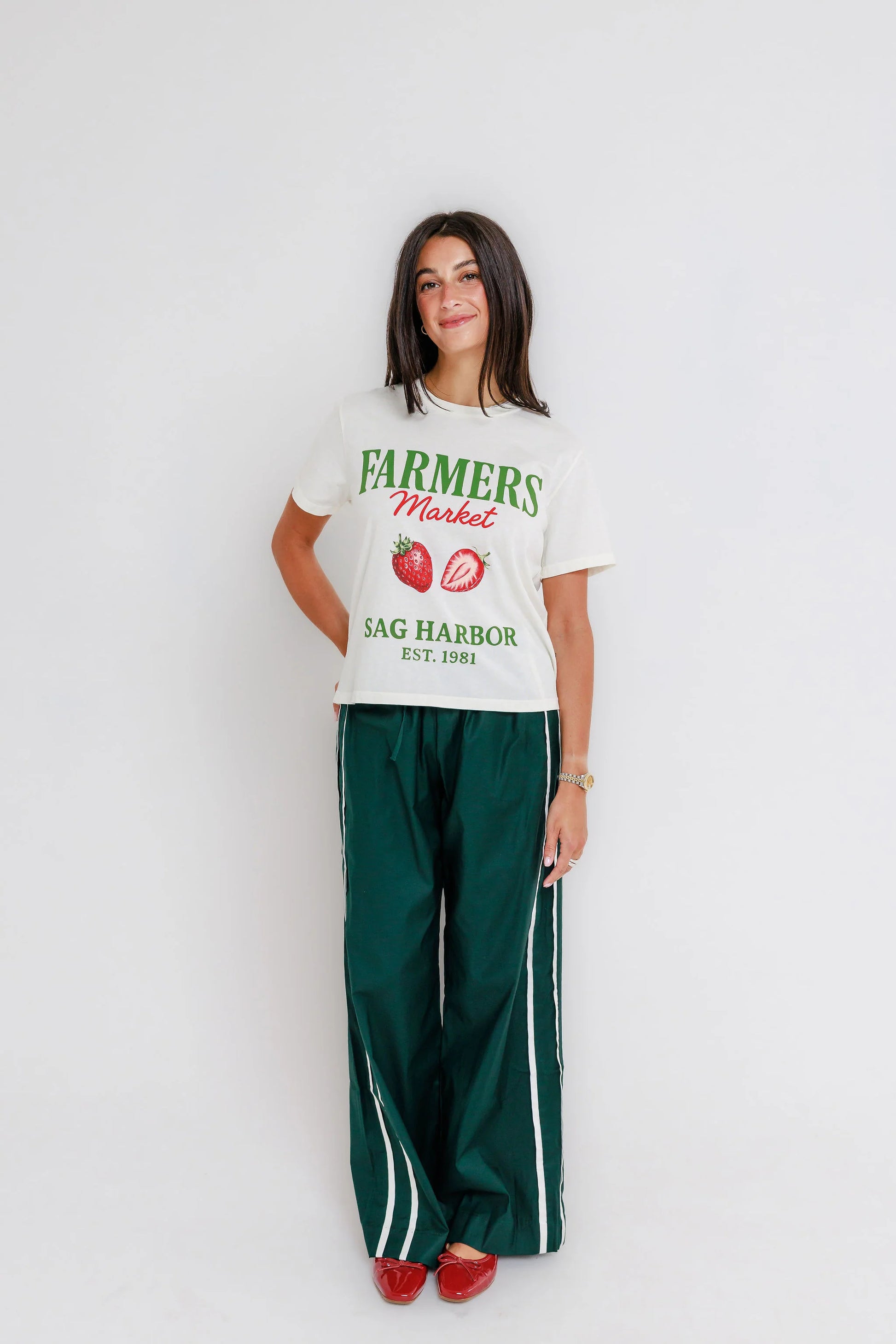 Woman in off-white cotton tee, green track pants, and red shoes standing against a white background.