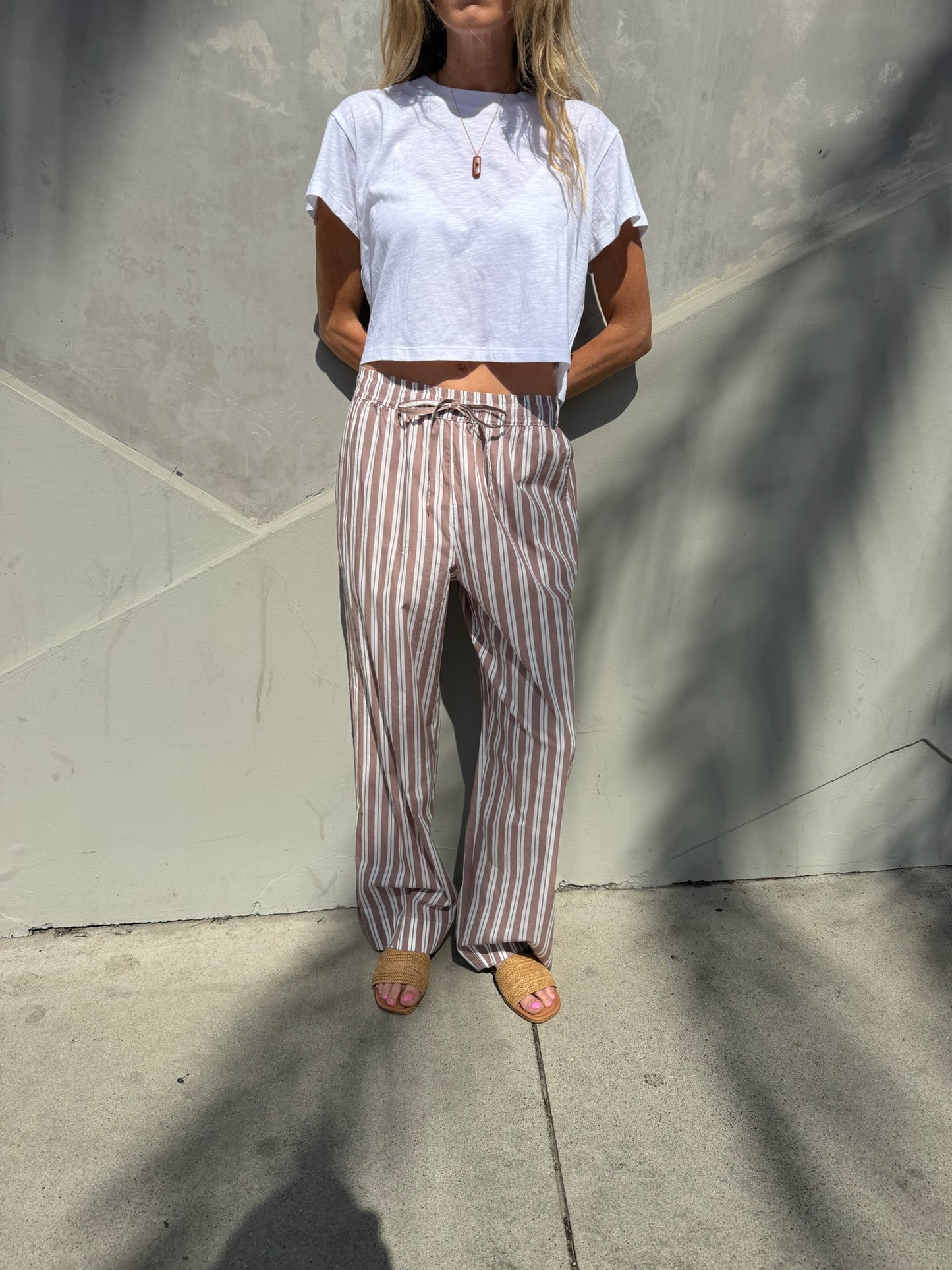 Woman wears wide-leg striped lounge pants in stucco, standing by a gray wall with sidewalk shadows. Cotton blend for everyday use.