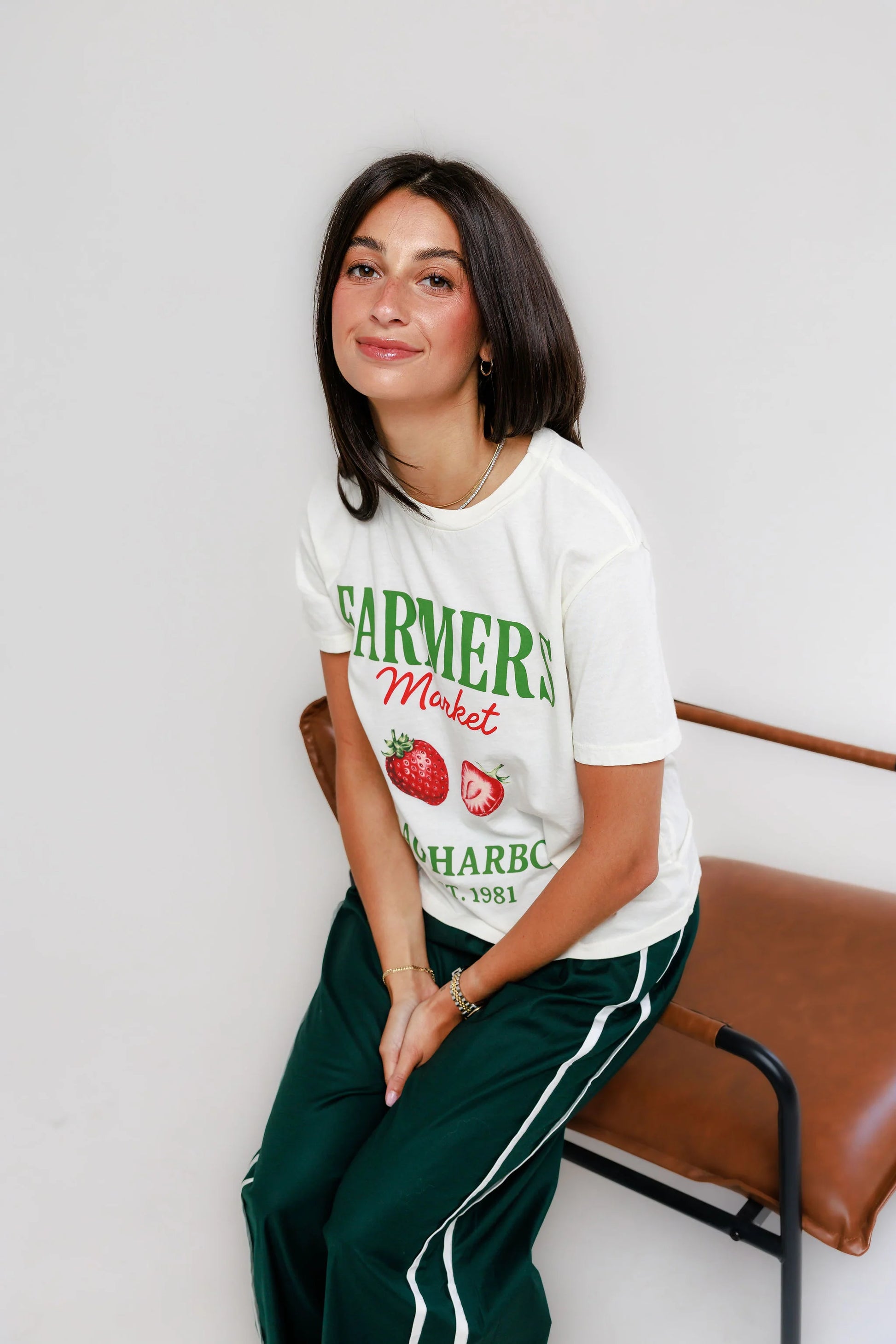 Woman with dark hair on a brown bench, wearing an off-white cotton tee and green pants suited for everyday use.