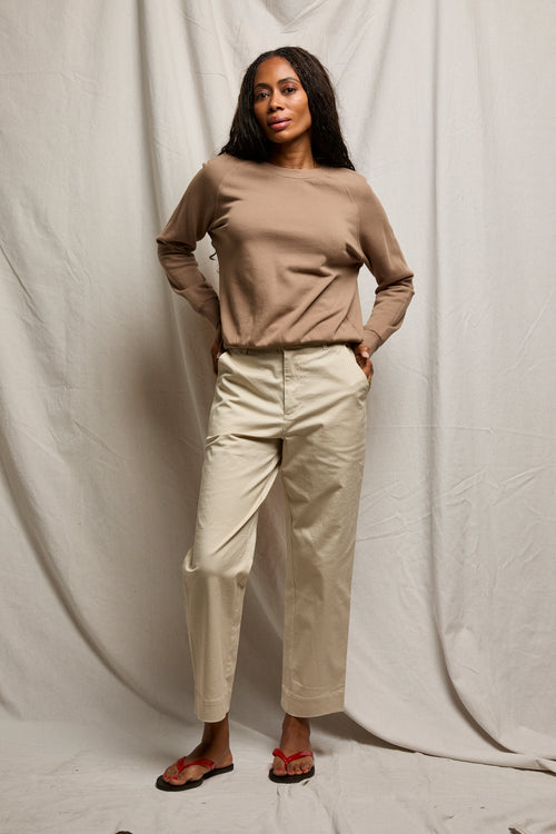 Woman in a white cotton sweatshirt, beige pants, and red sandals stands in front of a draped fabric backdrop.
