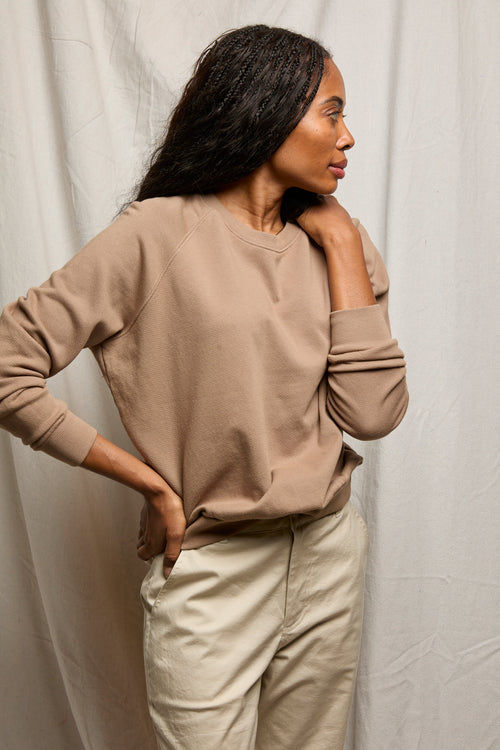 Woman in beige sweatshirt and light pants stands sideways with one hand on neck, highlighting casual loungewear.