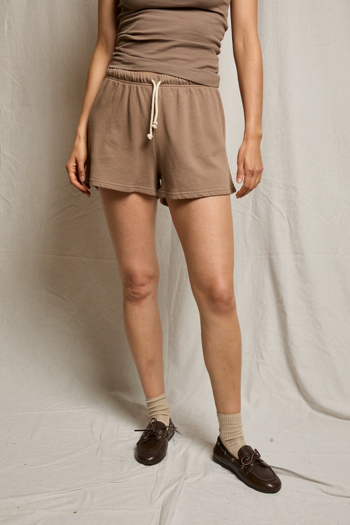 Person wears mocha cotton shorts, a brown tank top, tan socks, and loafers, standing before a neutral background.