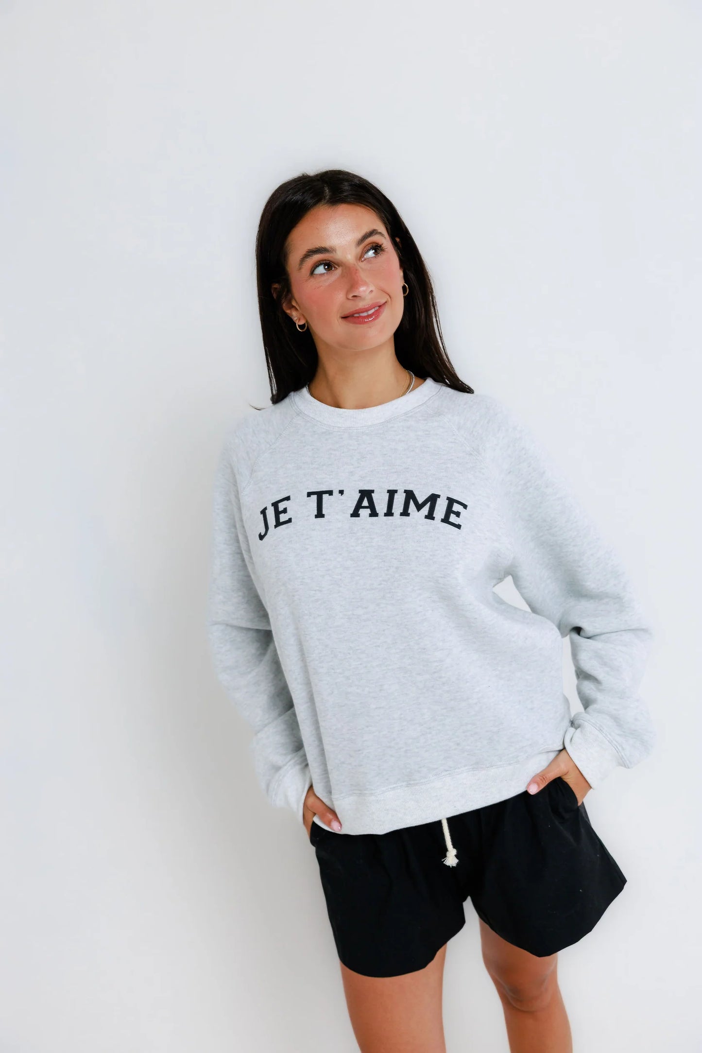 Woman wearing a light raglan crew sweatshirt and black shorts stands against a white background, looking to the side.