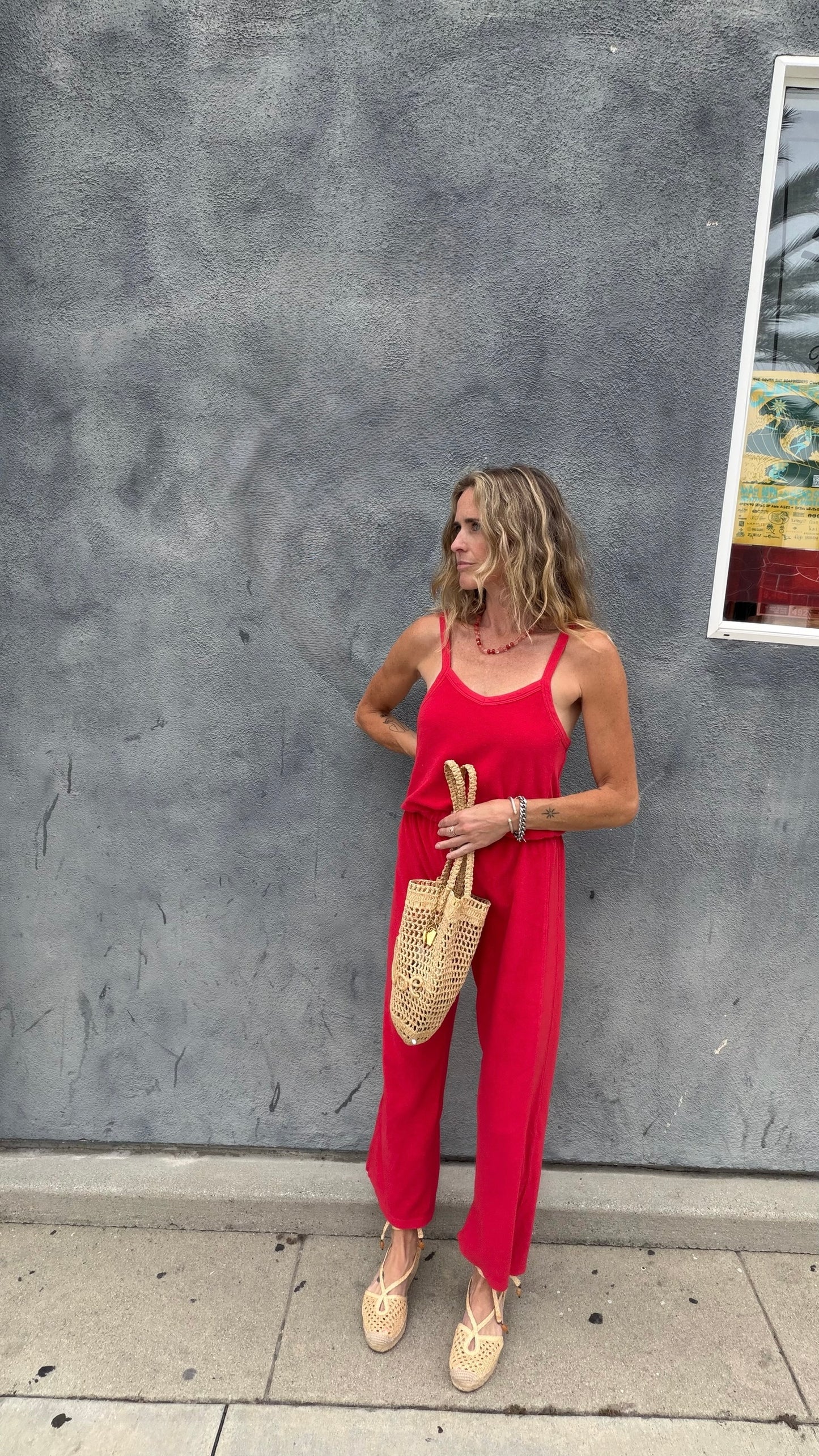 Woman holds woven bag, wearing cherry red jumpsuit, standing by window with gray textured wall in background.