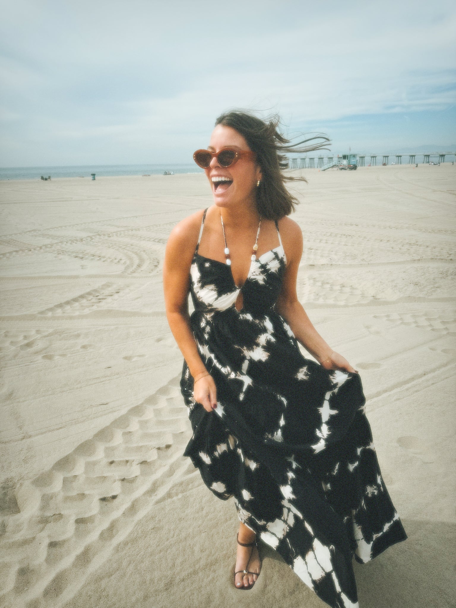 Woman on sandy beach in a white poplin dress; pier and tire tracks visible in the background.