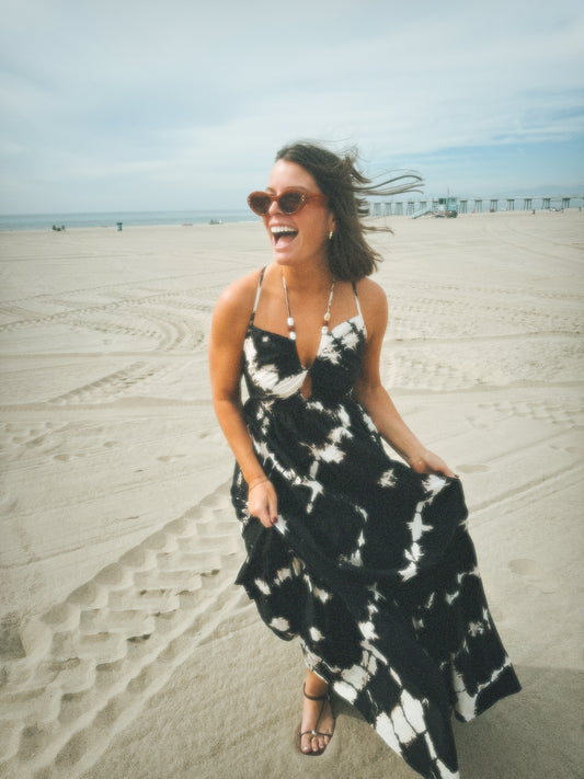 Woman on sandy beach in a white poplin dress; pier and tire tracks visible in the background.