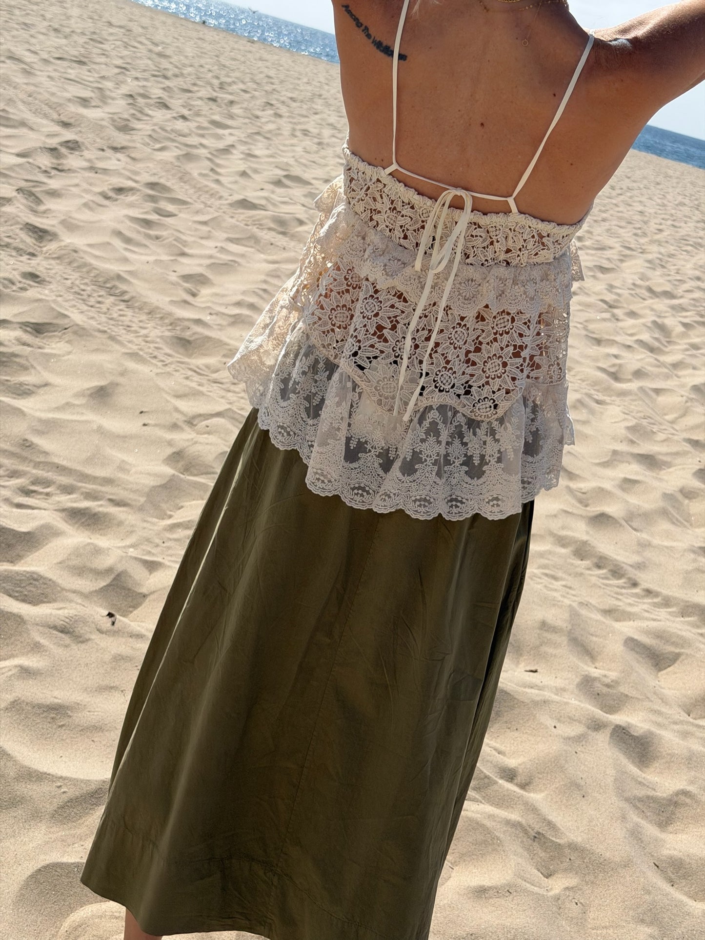 Woman in a lacy white top and dark olive midi skirt with arms raised, standing by the ocean.