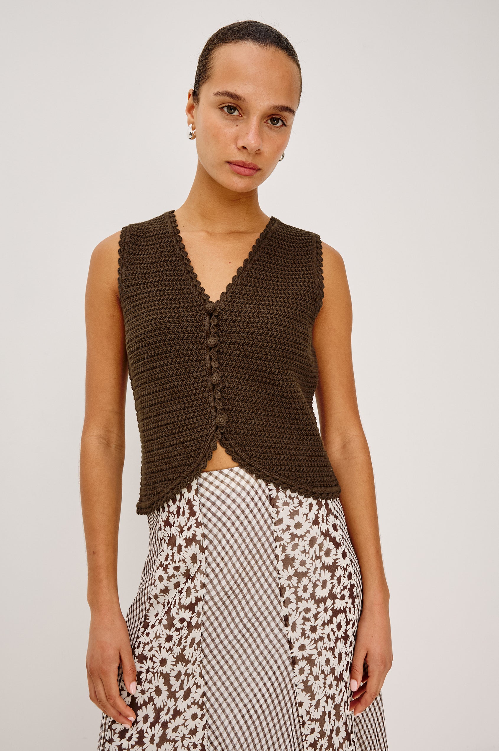 Woman in an espresso-colored vest and floral-checkered skirt stands against a plain background.