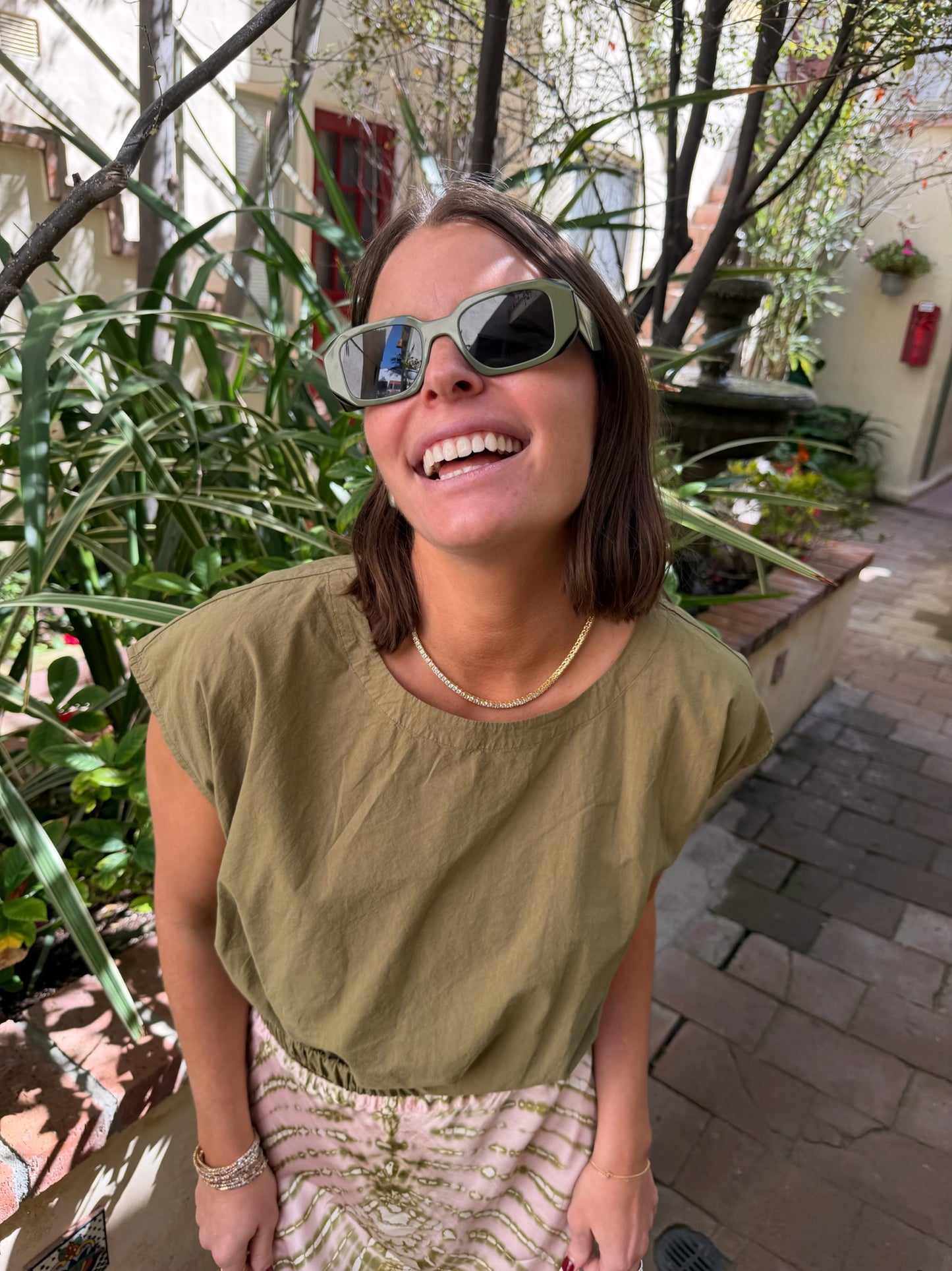 Woman in sunglasses wears a dark olive knit top, standing outdoors on a sunlit brick pathway.