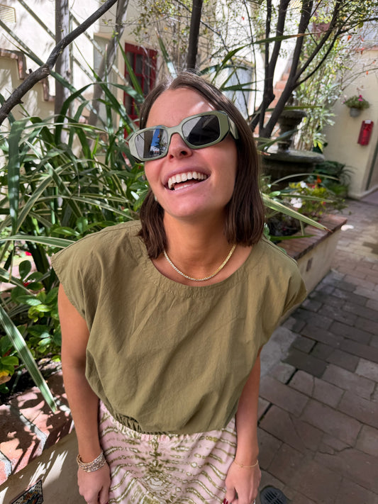 Woman in sunglasses wears a dark olive knit top, standing outdoors on a sunlit brick pathway.