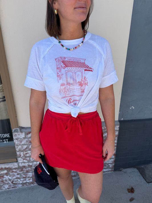 Woman in cream ringer crew tee and red skirt, holding a black hat while standing on the sidewalk.