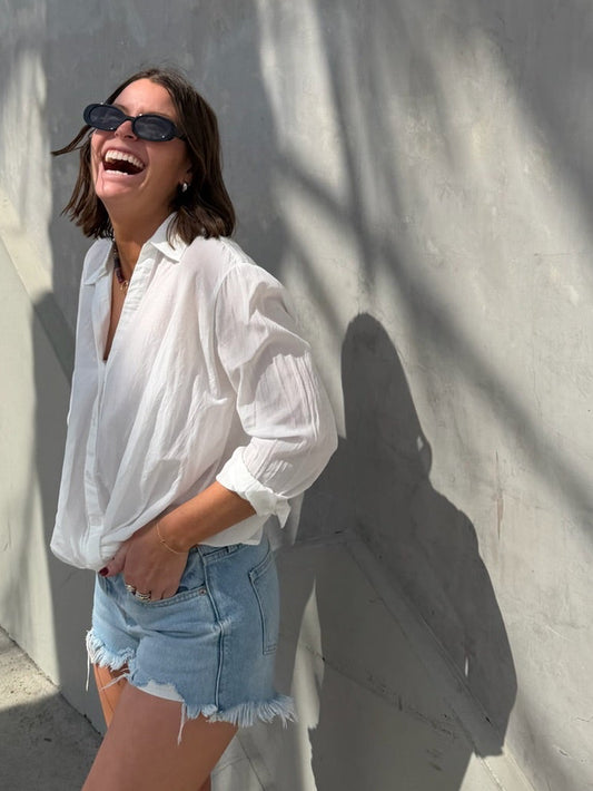 Woman in sunglasses smiles by a sunlit wall, wearing an off-white linen Traveler Top suited for casual wear.